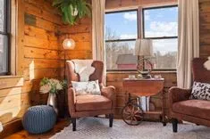 Cozy rustic cabin interior with two leather armchairs, wooden walls, a window with a countryside view, and a vintage side table with a lamp and decor.