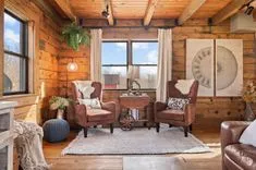 Cozy rustic living room interior with two brown leather armchairs, wooden walls and ceiling, area rug, decorative lamp, and a wagon wheel table, framed art on the wall, and sunlight streaming through the windows.