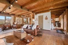 Cozy rustic living room with leather sofas, wooden floors and ceiling, and tasteful decorations, leading to a kitchen with a stairway on the side.