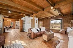 Spacious rustic living room with wooden beams, hardwood floors, leather furniture, and large windows overlooking a countryside view.
