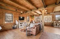Rustic wooden cabin interior with exposed beams, a cozy living area with leather sofas, and a fireplace under a mounted flat-screen TV.
