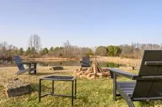 Outdoor relaxation area with Adirondack chairs, a fire pit with wood, and a small table overlooking a tranquil pond surrounded by trees.