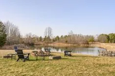 Outdoor relaxation area with Adirondack chairs around a fire pit by a tranquil pond, with bare trees in the background on a clear day.