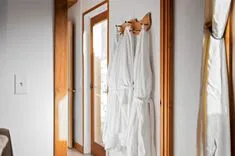 Two white bathrobes hanging on wooden hooks on a wall next to a mirror and a door.