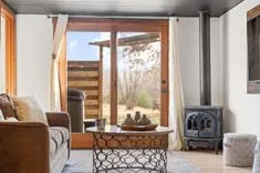Cozy living room interior with a couch, decorative metal coffee table holding vases, visible wood stove, opened French doors leading to a wooden deck, and sunlit curtains.