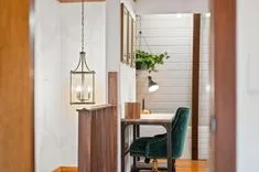 Elegant interior with a minimalist study area featuring a green upholstered chair with gold studs, a wooden desk with a lamp, and hanging indoor plants, all illuminated by a classic hanging pendant light.