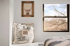 A cozy room corner with a decorative pillow on a window seat, a framed picture of a Highland cow on the wall, and a view of trees through the window.