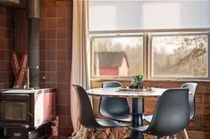 Cozy cabin interior with a wood stove, a dining table with chairs, and a view of a red barn through the window.