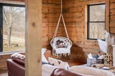 Cozy wooden cabin interior with hanging swing chair, plush cushions, and a view of outside trees through window.