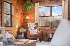 Cozy cabin interior with warm lighting featuring a leather armchair, hanging fern, large windows, and wooden walls.
