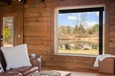 Cozy interior of a log cabin with a view of an outdoor seating area by a pond through a large window.
