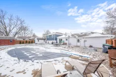 Backyard with snow-covered ground, patio furniture, a pool covered with a dark tarp, surrounded by a wooden fence, and houses in the background under a partly cloudy sky.