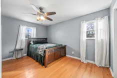 A well-lit bedroom with pale blue walls, hardwood floors, a ceiling fan with a light, a dark wood bed frame with green bedding, white curtains on a window, and a small black side table.