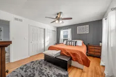 Modern bedroom interior with king-sized bed, burnt orange comforter, white and gray accent pillows, dark accent wall with artwork, ceiling fan, white doors, and a wooden dresser.