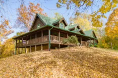 Wooden cabin with green roof surrounded by autumn foliage and a covered porch, set on a hill blanketed with fallen leaves.