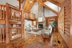 Spacious log cabin interior with high vaulted ceiling, stone fireplace, wooden staircase, and cozy living area with comfortable seating.