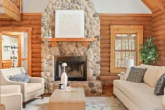 Cozy cabin living room with a stone fireplace, wooden log walls, comfortable seating, and natural light.
