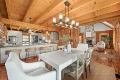 Spacious rustic log cabin interior with open plan design featuring a kitchen with modern appliances, a dining area with a large white table and chairs, and a living room with a stone fireplace. Wooden beams and hardwood floors throughout.