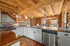 Interior of a rustic wooden kitchen with stainless steel appliances, white cabinets, and log cabin walls with pendant lighting.