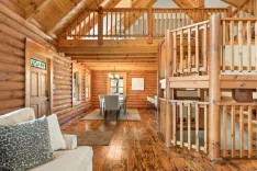 Interior of a spacious log cabin with exposed wooden beams, hardwood floors, and a loft area with railing.