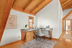 A bright home office space with a wooden desk, upholstered chair, and an abstract wall art piece set against natural wood beams and flooring.
