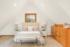 Bright and airy attic bedroom with a queen-size bed, white bedding, wooden dresser, and angled ceilings with recessed lighting.