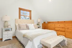 Cozy bedroom interior with a queen-sized bed, white and tan bedding, a knitted throw, natural wood furniture, and a bright window.