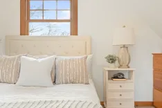 Cozy bedroom interior with beige upholstered headboard, decorative pillows, white bedside table with lamp and plant, and a view of trees through the window.