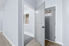 Bright, modern hallway with an open white door leading to a room with a visible window and a closed gray door at the end.