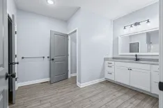 A clean and modern bathroom interior with a white vanity, granite countertop, and large mirror, featuring wooden flooring and a view into a connecting room.