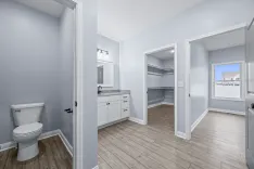 Interior view of a clean, empty bathroom connected to a walk-in closet with built-in shelves, featuring modern fixtures, white cabinetry, and light wood flooring.