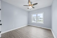 Empty room with light gray walls, wood flooring, and a ceiling fan, featuring a large window with a view of the neighborhood.