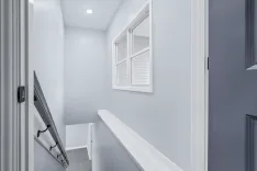 A well-lit, narrow corridor with gray walls, white trim, a small window with blinds, and a coat rack mounted on the wall.
