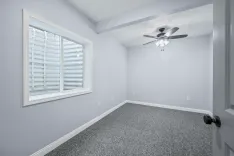 Empty room with gray walls, a large window with blinds, textured gray carpet, and a ceiling fan with lights.
