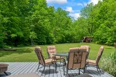 Outdoor patio area with a set of plush chairs around a glass-top table overlooking a lush green lawn and dense tree line with a small red shed in the background.