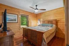 Cozy wooden cabin bedroom interior with a large bed, quilt bedding, hardwood floor, ceiling fan, and a scenic painting on the wall, illuminated by natural light from a window with greenery outside.