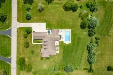 Aerial view of a suburban house with a swimming pool and a well-manicured lawn, surrounded by trees.