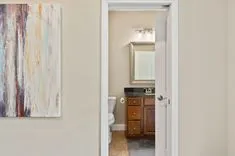 View from a hallway into a bathroom with wooden vanity and mirror, wall art on the left side.
