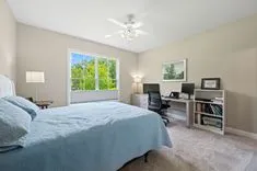Bright and airy bedroom interior with a bed covered in blue bedding, a home office desk with a computer by the window, bookshelves, and a ceiling fan.