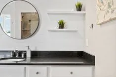 Modern bathroom interior with black countertop, white cabinets, round mirror, and decorative plants on shelves.