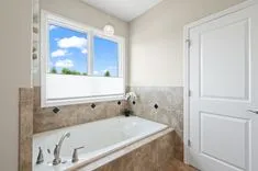 Bright bathroom interior with a built-in bathtub, tiled walls, a large window with a view of blue sky and clouds, and a closed white door.