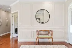 Elegant interior room with white wainscoting walls, a round mirror, a wooden bench, and a patterned area rug over hardwood floors.