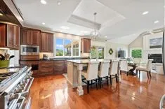 Spacious kitchen with rich wooden cabinetry, stainless steel appliances, a large island with bar stools, and a dining area in the background.
