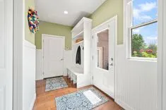 Bright and airy entryway with green walls, white trim, decorative coat hooks, a bench with storage, a colorful wall ornament, and natural light coming through a window.