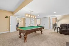 Spacious game room with a green-felt pool table, modern hanging light fixture, beige carpet, and cozy seating area.