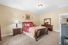 Cozy bedroom interior with a queen-sized bed adorned with red and beige bedding, matching wooden nightstand and dresser, a table lamp, wall art, plush carpeting, and a ceiling light fixture.