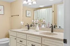 Modern bathroom interior with a beige color scheme, featuring a double sink vanity with granite countertop, framed flower art on the walls, and a shower curtain with floral print.