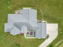 Aerial view of a large residential house with a complex roof design, surrounded by a well-manicured lawn, a patio area with a pergola, and a driveway leading to an attached garage.