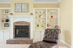 Cozy living room corner with a brown leather armchair, white built-in bookshelves decorated with various ornaments, and a fireplace with a brick surround.