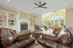 Spacious living room with leather sofas, a central fireplace, built-in shelving with various decorative items, large arched windows, and a sunroom extension visible in the background.